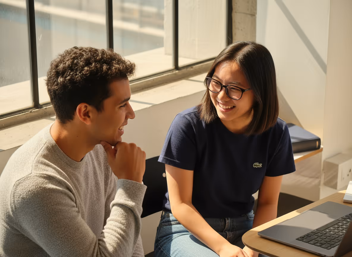 Two young people smiling and talking while sitting by a table with a laptop in a sunlit room.